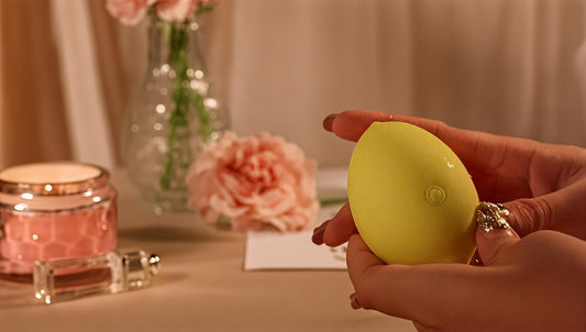 Hand holding yellow egg-shaped adult toy with pink flowers and candle in background