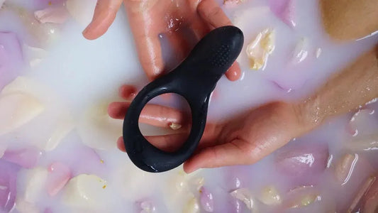 Black silicone cock ring held in hands over milky bath with flower petals